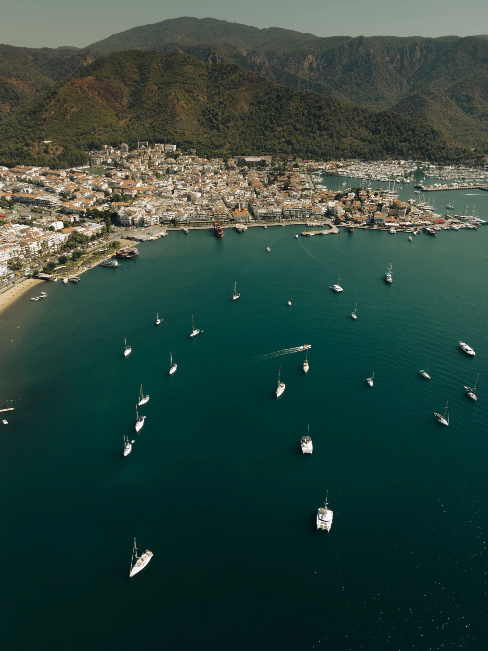 A stunning aerial view of a coastal town with yachts anchored in the harbor and lush mountains in the background.